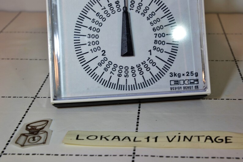 May include: White kitchen scale with a black dial and needle. The dial has markings for grams and kilograms. The scale reads 3kg x 25g. The text "EKS DESIGN OCHOT SK" is printed on the scale.