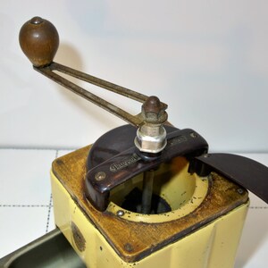 May include: A vintage yellow and brown coffee grinder with a wooden handle and a black plastic lid. The grinder has a metal crank and a metal base. The lid is open and the grinder is ready to be used.