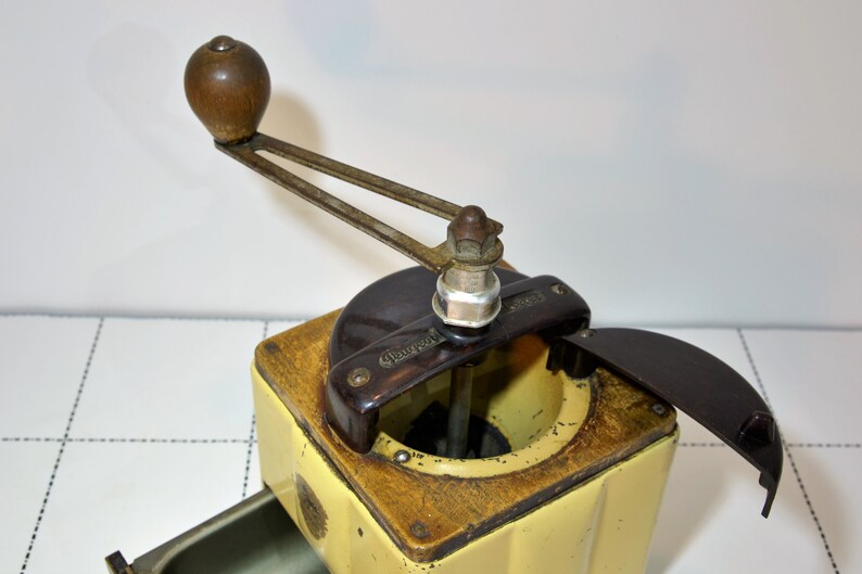 May include: A vintage yellow and brown coffee grinder with a wooden handle and a black plastic lid. The grinder has a metal crank and a metal base. The lid is open and the grinder is ready to be used.