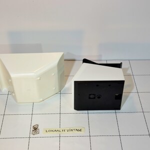 May include: Two white plastic containers with black accents. One container is open and the other is closed. The containers are on a white and black checkered surface.