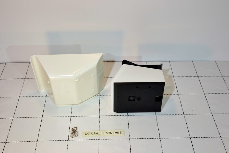 May include: Two white plastic containers with black accents. One container is open and the other is closed. The containers are on a white and black checkered surface.