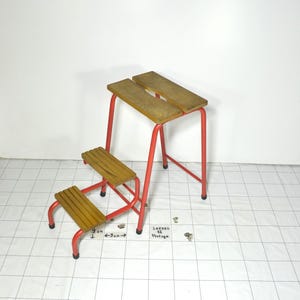 Puede incluir: Un taburete vintage con estructura de metal rojo y escalones de madera. El taburete más pequeño tiene dos escalones, y el más grande tiene dos tablones de madera. Los escalones miden aproximadamente 9 cm de ancho. También incluye el texto "Logees 11 Vintage".