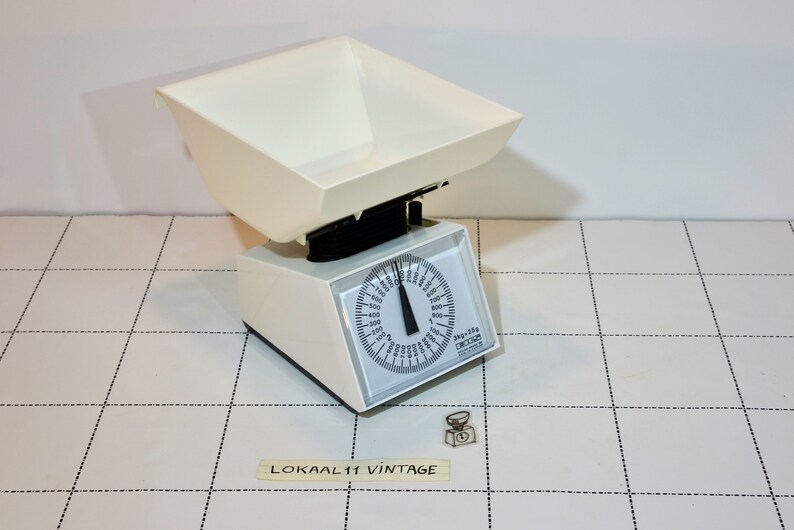 May include: White vintage kitchen scale with a large bowl and a round dial. The dial has markings for grams and kilograms. The scale is on a white tiled surface.