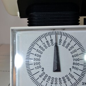 May include: A white kitchen scale with a black dial and a black needle. The dial has numbers from 100 to 900 in increments of 100. The needle is pointing to 0. The scale has a label that says "3kg x 25g".