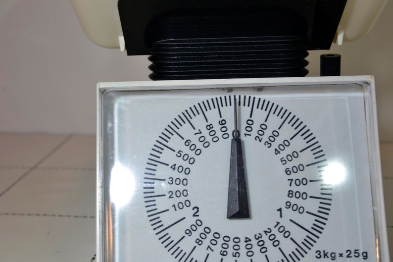 May include: A white kitchen scale with a black dial and a black needle. The dial has numbers from 100 to 900 in increments of 100. The needle is pointing to 0. The scale has a label that says "3kg x 25g".