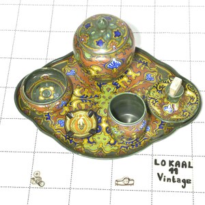 Può includere: Un set da toeletta in ceramica composto da un vassoio, un barattolo con coperchio, una ciotolina, una tazza e un piattino con un perno centrale. I pezzi sono decorati con un vivace motivo multicolore nei toni del verde, arancione, blu e giallo. Testo LOKAAL 11 Vintage.