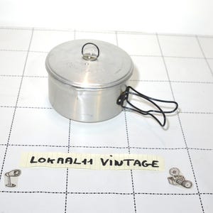 Può includere: Una pentola da cucina vintage in alluminio con coperchio e manico in filo di ferro nero. La pentola è cilindrica e ha un piccolo anello sul coperchio. Il testo "LOKRAAL11 VINTAGE" è su un pezzo di carta sotto.