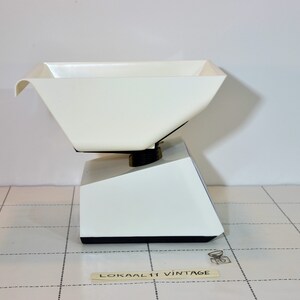 May include: White kitchen scale with a white plastic bowl. The scale has a black base and a black knob on the side. The scale is sitting on a white tiled surface.
