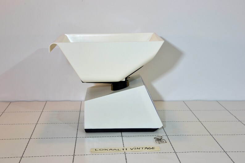 May include: White kitchen scale with a white plastic bowl. The scale has a black base and a black knob on the side. The scale is sitting on a white tiled surface.