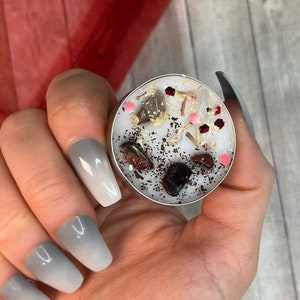 May include: A white tea light candle with embedded crystals, including clear quartz, amethyst, rose quartz, and black obsidian. The candle is surrounded by glitter and is in a person's hand.