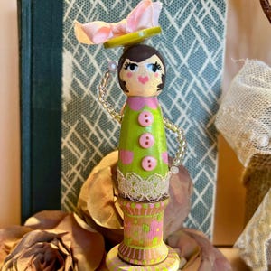 May include: A whimsical wooden doll with a green and pink dress, a pink bow, and a green hat. The doll is standing on a painted wooden spool.