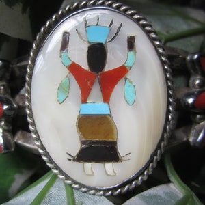 May include: A silver bracelet with a round, white mother-of-pearl inlay featuring a colorful Native American figure. The figure is surrounded by turquoise, red, and black stones.