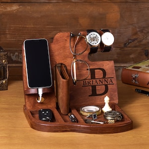 Puede incluir: Un organizador de madera con compartimentos para un teléfono, una cartera, un reloj, gafas y otros objetos pequeños. El organizador está personalizado con el nombre "BRIANNA" en una fuente decorativa.