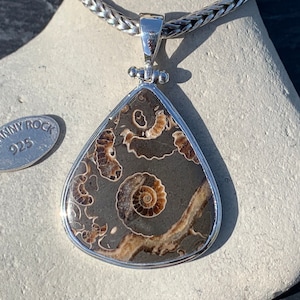 May include: A silver pendant necklace featuring a teardrop-shaped stone with a brown and white ammonite fossil pattern. The pendant is attached to a silver chain with a braided design. The tag attached to the chain reads "Johnny Rock 925".