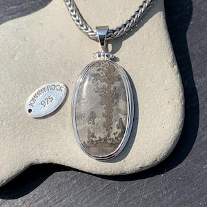 May include: Oval pendant with a silver chain. The pendant features a gray and brown stone with a tree-like pattern, set in a silver frame. A small silver tag with the text "JOHNNY ROCK 925" is next to the pendant.