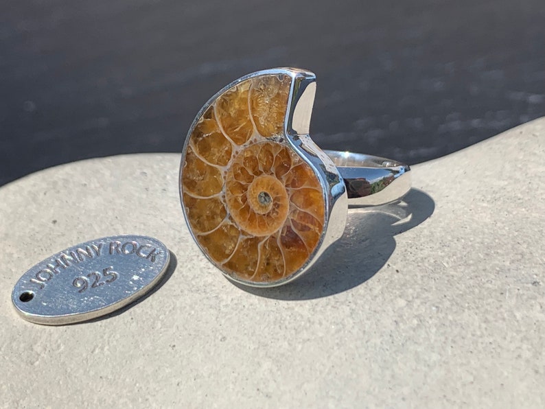 May include: A silver ring with a large ammonite fossil set in the center. The ring is on a gray surface with a small silver tag that says "Johnny Rock 925".