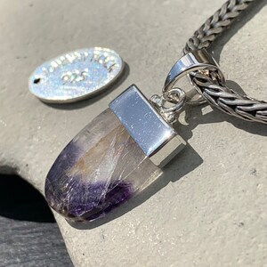 May include: A silver pendant with a purple and white crystal stone. The pendant is attached to a silver chain.