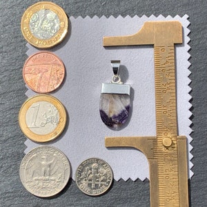 May include: A silver pendant with a purple and white fluorite stone. The pendant is approximately 2.5 cm long and is shown next to a ruler and various coins for size comparison.