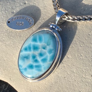 May include: A silver pendant with a blue and white gemstone set in a silver oval frame. The pendant is attached to a silver chain. The tag attached to the chain reads "Johnny Rock 925".