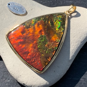May include: A triangular pendant with a gold setting and a large, iridescent gemstone. The gemstone has shades of red, orange, green, and gold. The pendant is on a white rock.