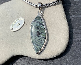 Puede incluir: Un colgante de plata con una piedra verde y negra. La piedra tiene un patrón de remolino. El colgante está en una cadena de plata. La etiqueta dice "Johnny Rock 925".