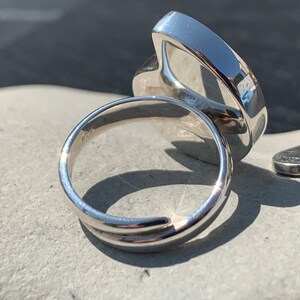 May include: A silver ring with a large, smooth, oval-shaped stone. The ring is shown from the side, with the stone facing away from the camera. The ring is sitting on a gray surface.