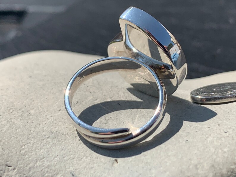 May include: A silver ring with a large, smooth, oval-shaped stone. The ring is shown from the side, with the stone facing away from the camera. The ring is sitting on a gray surface.