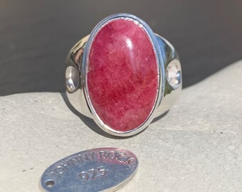 Pode incluir: Um anel de prata com uma grande pedra oval vermelha engastada no centro. O anel tem uma banda de prata com dois pequenos furos de cada lado da pedra. O anel está sobre uma superfície branca com uma etiqueta de prata que diz "Johnny Rock 925" nela.