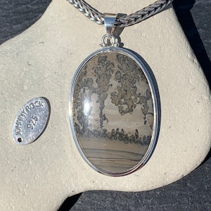 May include: A silver pendant with a large oval-shaped stone featuring a brown and gray dendritic agate pattern. The pendant is on a silver chain and has a small silver tag with the text "JOHNNY ROCK 925" on it.