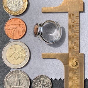 Puede incluir: Un anillo de plata con una piedra marr&oacute;n engastada en la banda. El anillo se est&aacute; midiendo con una regla, que muestra que el anillo tiene un di&aacute;metro de aproximadamente 2,5 cm.