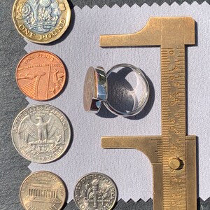 May include: A silver ring with a dark gray stone set in a silver bezel. The ring is being measured with a brass caliper. The ring is surrounded by various coins, including a British one pound coin, a British one penny coin, and three US coins: a quarter, a dime, and a penny.