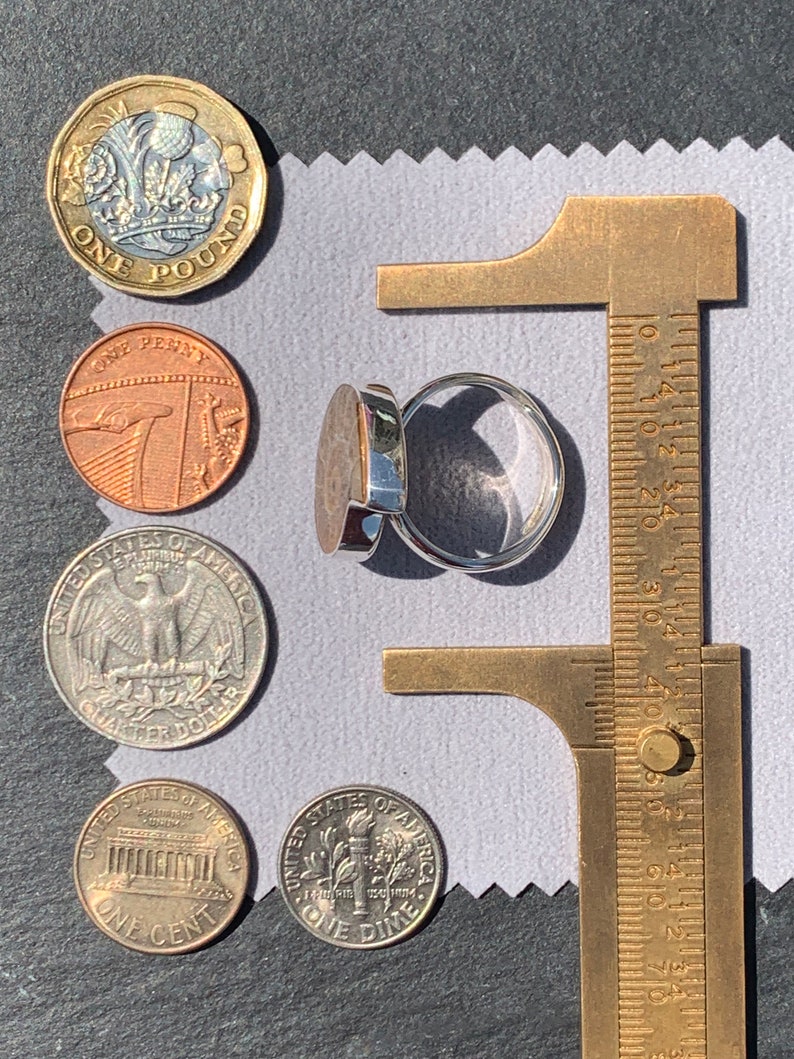 May include: A silver ring with a dark gray stone set in a silver bezel. The ring is being measured with a brass caliper. The ring is surrounded by various coins, including a British one pound coin, a British one penny coin, and three US coins: a quarter, a dime, and a penny.