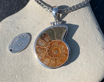 Puede incluir: Un collar de plata con un colgante que presenta un amonite fosilizado. El colgante está engastado en un marco de plata y tiene un patrón de espiral marrón y blanco. El collar está en una cadena de plata trenzada. La etiqueta adjunta al collar dice "Johnny Rock 925".