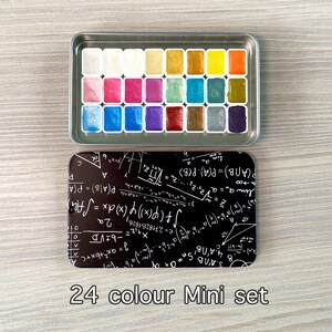 May include: A 24-color watercolor paint set in a silver metal case. The paint pans are arranged in a grid pattern and are filled with various colors, including gold, silver, blue, green, and purple. The case has a black background with white mathematical equations and formulas.