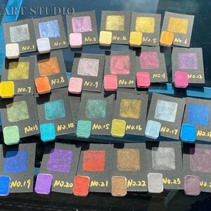 May include: A collection of 24 small square paint samples, each labeled with a number from 1 to 24. The samples are arranged in rows on black cards. The paint colors range from metallic gold to blue, green, purple, and red.