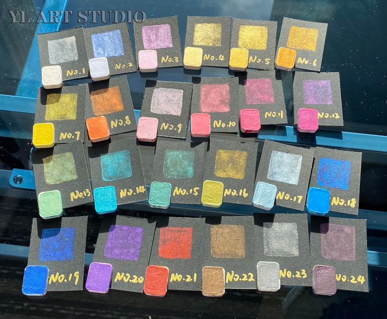 May include: A collection of 24 small square paint samples, each labeled with a number from 1 to 24. The samples are arranged in rows on black cards. The paint colors range from metallic gold to blue, green, purple, and red.