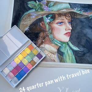 May include: A set of 24 watercolor paints in a travel box. The paints are arranged in a grid of 4 rows and 6 columns. The paints are a variety of colors, including silver, gold, pink, blue, green, and purple. The box is white with a silver trim. The text "24 quarter pan with travel box" is printed on the bottom of the image. The text "YL art" is printed in a cursive font on the bottom right of the image.