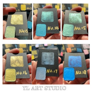 May include: Close-up of ten small square paint samples on black cards. The samples are arranged in two rows of five. The samples are numbered 13 through 18. The paint colors are gold, green, blue, and teal.