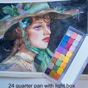 May include: A watercolor painting of a woman wearing a large, green hat with flowers. The painting is next to a 24-pan watercolor set with a light box.