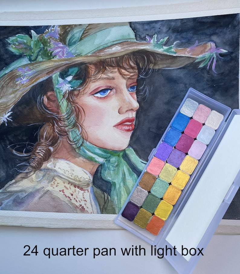 May include: A watercolor painting of a woman wearing a large, green hat with flowers. The painting is next to a 24-pan watercolor set with a light box.