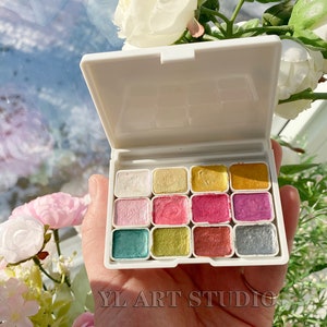 May include: A white plastic palette with 12 small, square paint pans. The pans are filled with different colors of metallic paint, including gold, silver, copper, and pink.