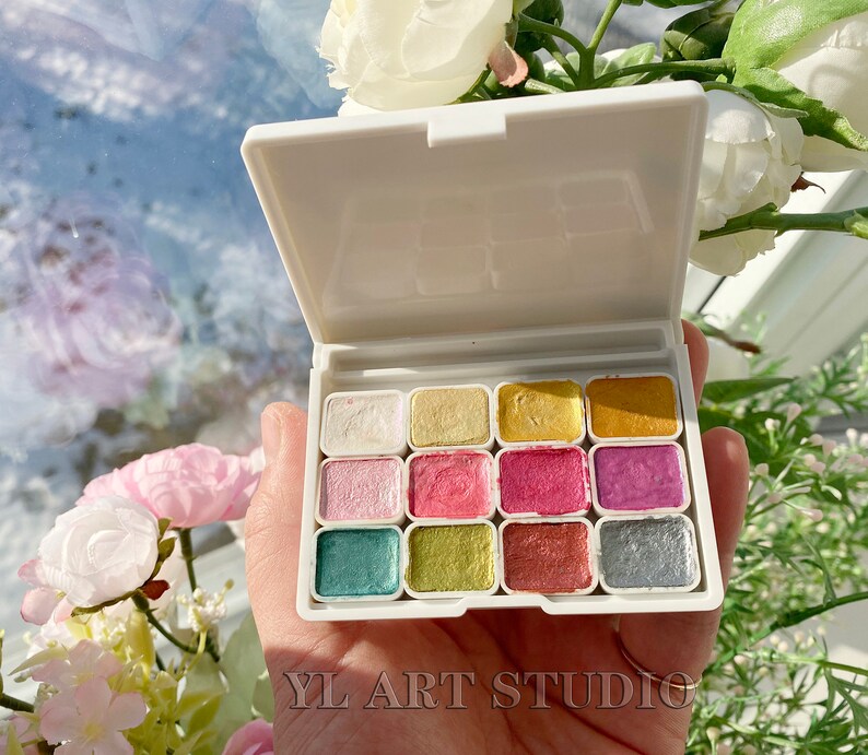 May include: A white plastic palette with 12 small, square paint pans. The pans are filled with different colors of metallic paint, including gold, silver, copper, and pink.