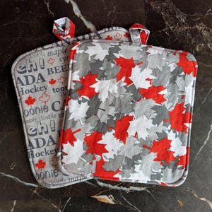 May include: Two square pot holders. One is gray with red maple leaves. The other is gray with the words "Canada", "eh!", "Hockey", and "toonie" with red maple leaf accents. Both have a loop for hanging.