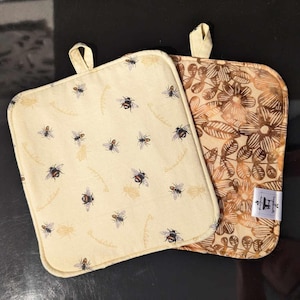 Puede incluir: Dos agarraderas de olla beige. Una tiene un patrón floral y la otra tiene un patrón de abejas y flores. La agarradera floral tiene un esquema de color marrón y beige. La agarradera de abejas tiene un fondo beige con abejas y flores marrones. Ambas agarraderas tienen un borde cosido marrón.