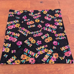 Può includere: Sottobicchiere in tessuto nero con un motivo floreale colorato. Il motivo presenta fiori rosa, arancioni, gialli e turchesi su uno sfondo nero.