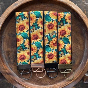 May include: Four key fobs featuring a sunflower and butterfly design. The fabric is yellow with orange sunflowers, green leaves, and butterflies. Each fob has a different metal clasp: bronze, rose gold, black, and olive green.