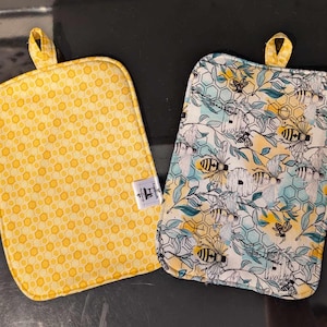 Peut inclure: Deux maniques jaunes et blanches avec un motif en nid d'abeille et des abeilles. Les maniques sont en tissu et ont un bord cousu.