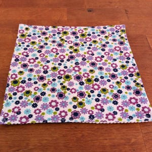 Può includere: Un quadrato di tessuto bianco con un motivo floreale in tonalità di viola, blu e verde. Il tessuto ha un bordo cucito.