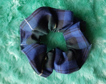 May include: A blue, black, and white plaid scrunchie. The fabric is a classic tartan pattern, with a mix of dark blue, black, and white stripes. The scrunchie is gathered and circular, designed to hold hair.