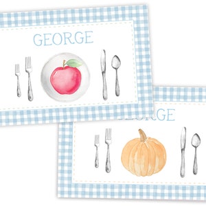 Op de afbeelding: Twee blauwe en witte gingham placemats met een witte rand. De bovenste placemat heeft een rode appel op een wit bord met een vork, mes en lepel. De onderste placemat heeft een oranje pompoen met een vork, mes en lepel. Beide placemats hebben de naam "GEORGE" erop gedrukt.